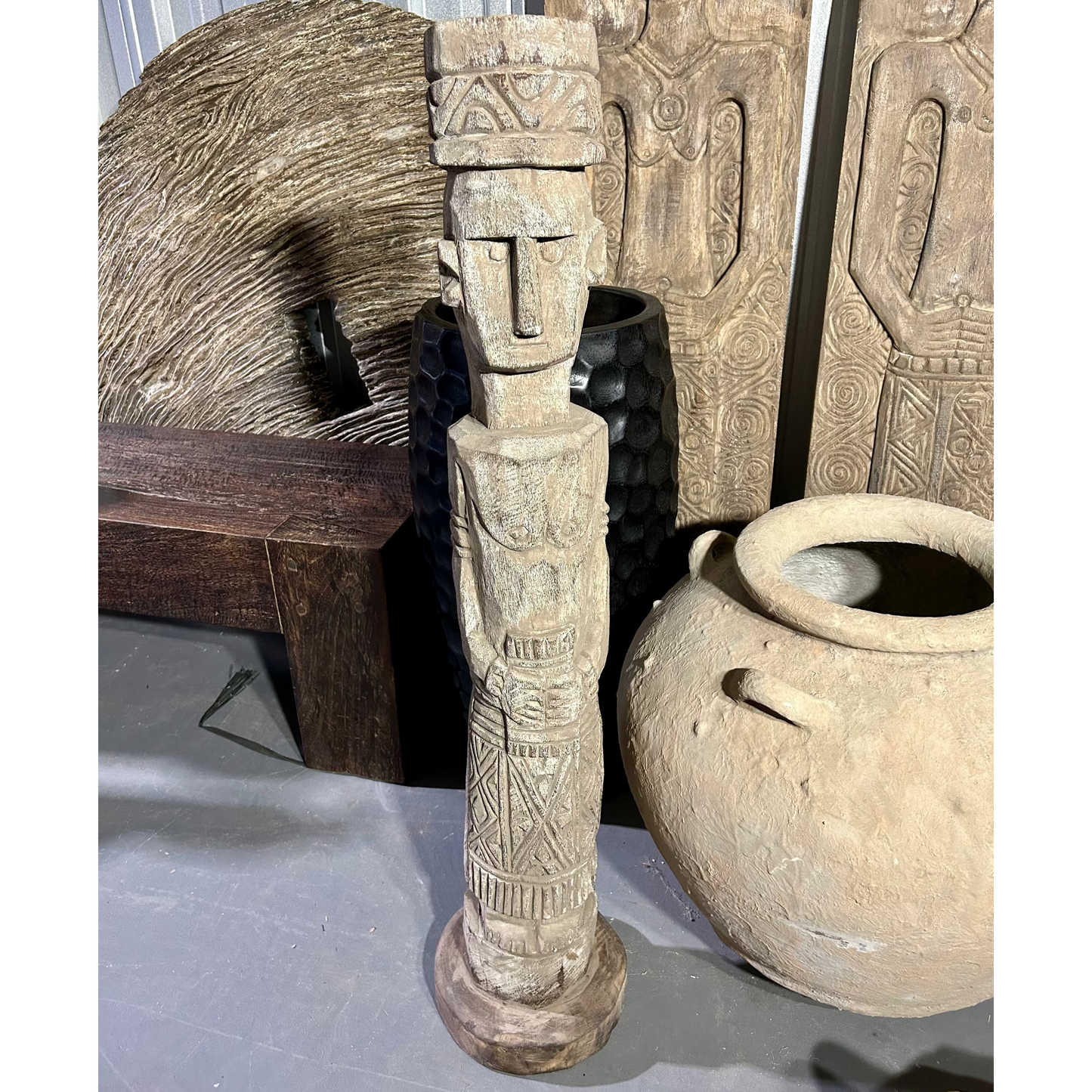 A Female Timor Ancestor statue with a human-like face and body, topped with a stylized, textured hat, displayed among other art pieces.