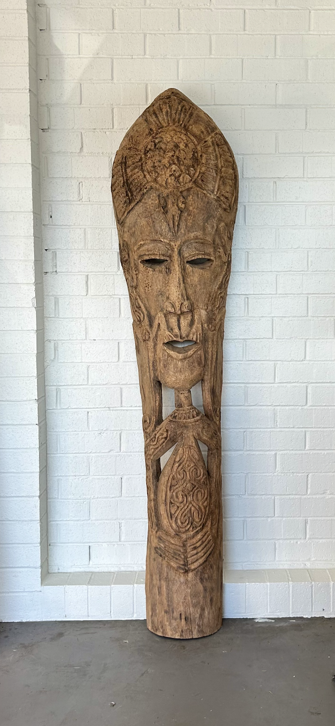 A tall, slender hand-carved coconut wood totem with a distinctive fibrous grain and a tribal-inspired face against a white painted brick background.