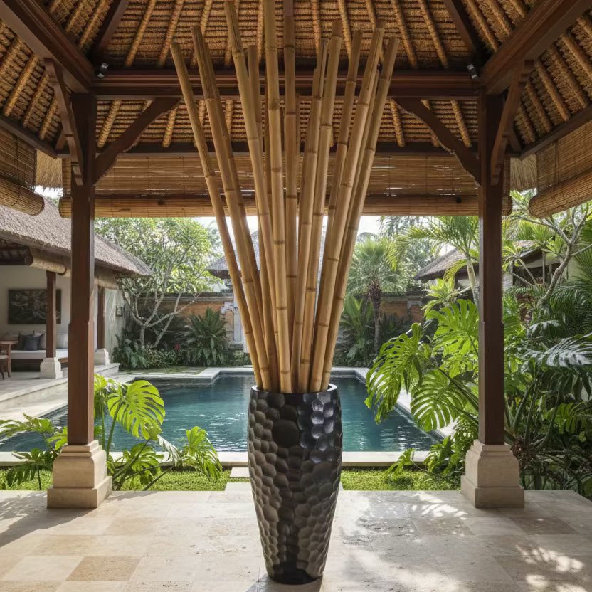 A lifestyle image of a tall, black, hand carved wooden pot filled with bamboo poles is placed on a stone paved floor under a thatched Balinese pavilion roof. An inground pool in the background surrounded by lush plants and an outdoor living space.