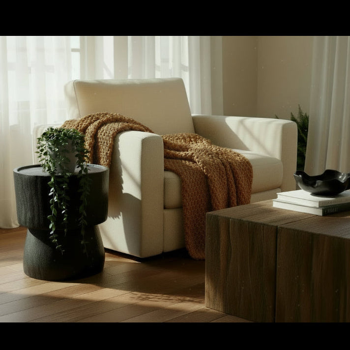 A round black timber side table next to a cream square single sofa with a trailing plant on top.