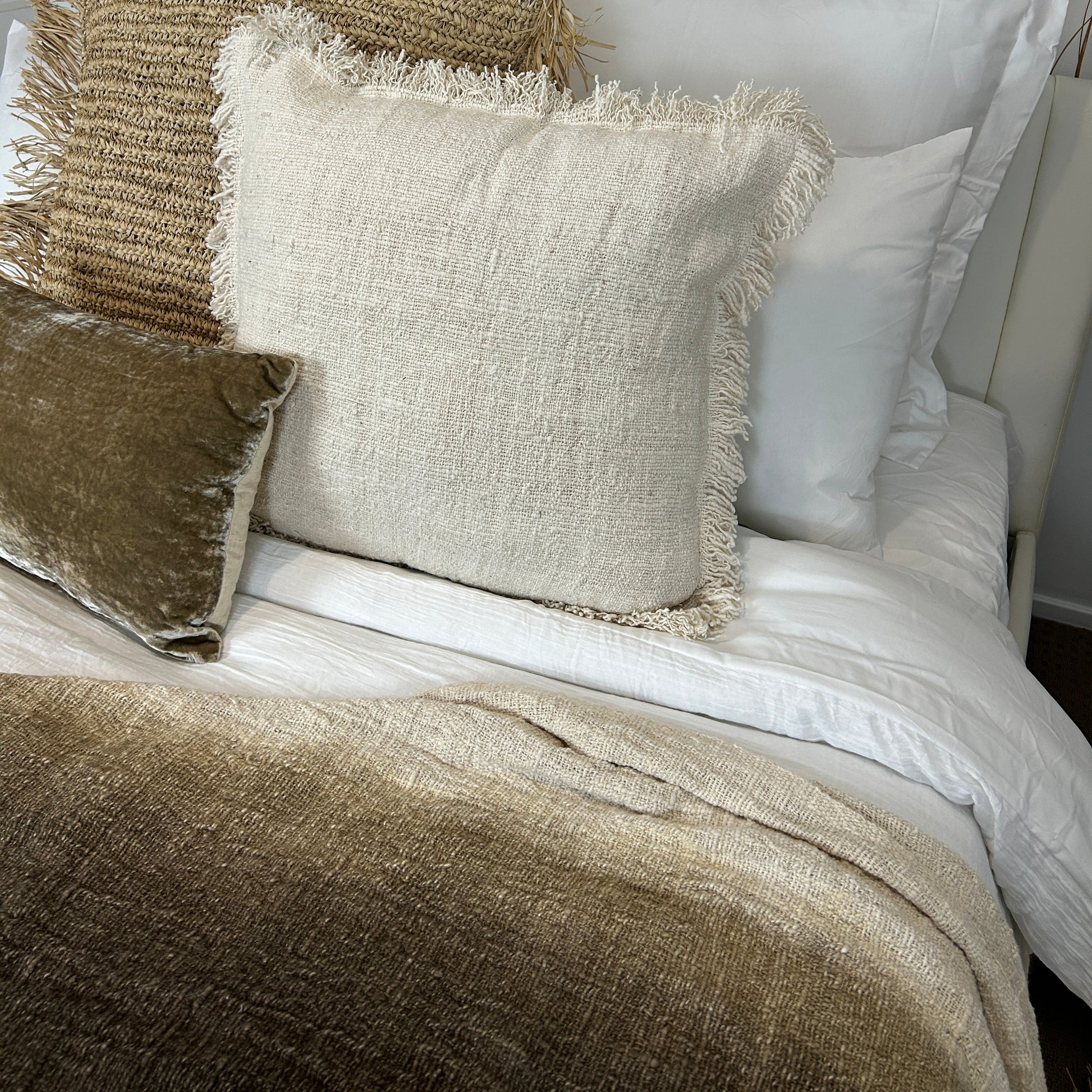 Espresso graduated throw is displayed on a bed featuring a natural woven cotton cushion, raffia cushion and velvet cushion against white bedding.