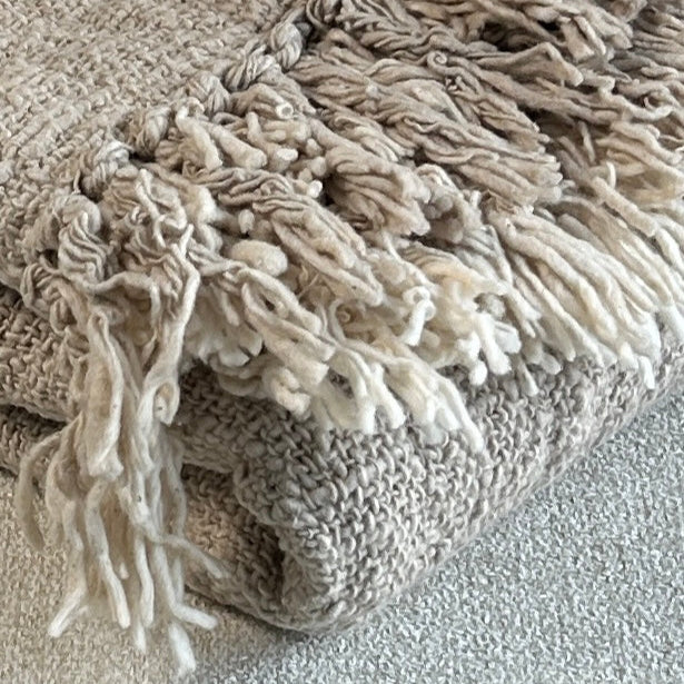 Folded cotton throw shown with fringe and woven detail close up. Latte colour of the throw set against a neutral fabric surface to highlight the soft and relaxed tones of beige.