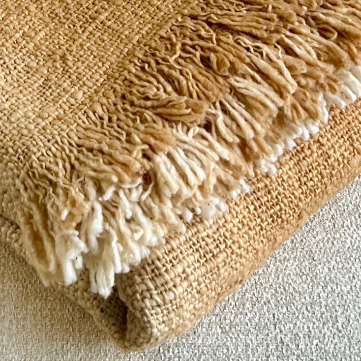 Folded cotton throw shown with fringe and woven detail close up. Golden spice colour of the throw set against a neutral fabric surface to highlight the golden tones of colour.