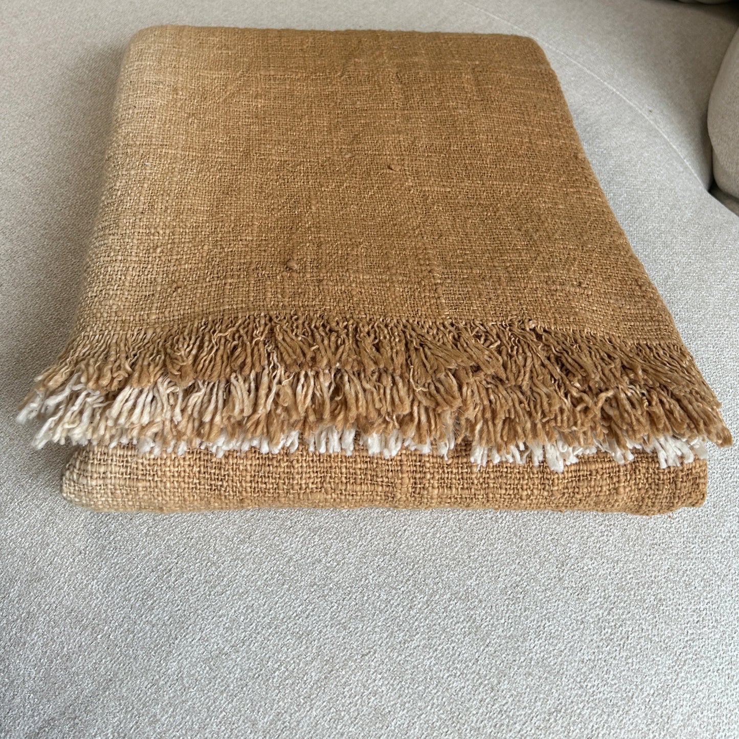 Full product image of folded cotton throw shown with fringe and woven detail close up. Golden spice colour of the throw set against a neutral fabric surface to highlight the golden tones of colour.