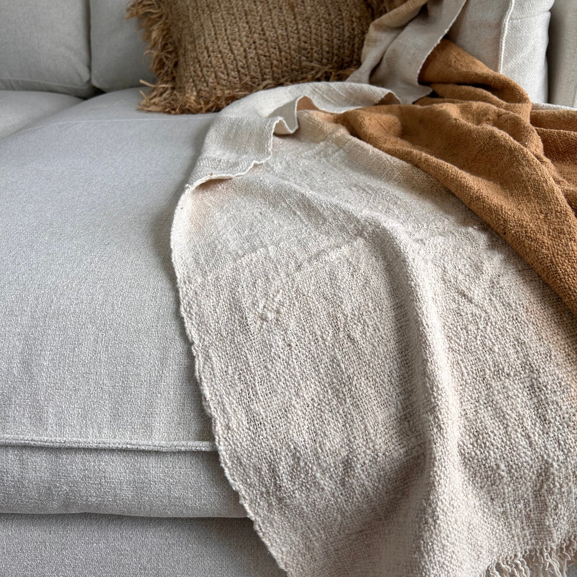 Lifestyle image of Spice cotton throw shown layered on a modular lounge paired with raffia cushion close up. Golden spice colour of the throw set against a neutral fabric surface to highlight the golden tones of colour.