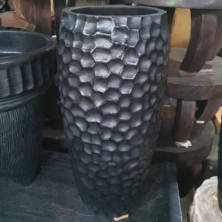 Handcarved tall black pot sitting next to a black side table with brown carved stools in the background. This photo was taken in the factory before shipping.