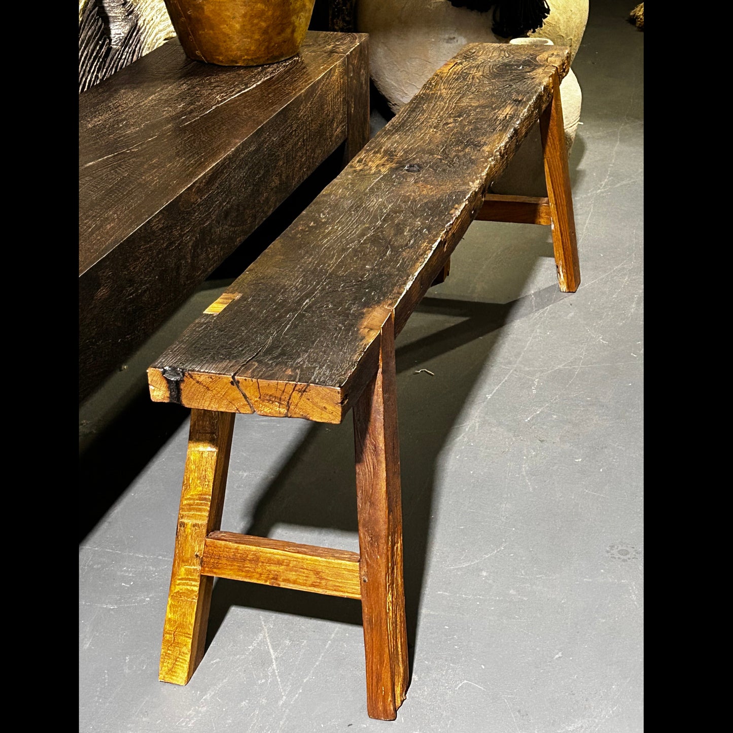 A vintage teakwood bench seat with a weathered patina displayed in an indoor warehouse setting.
