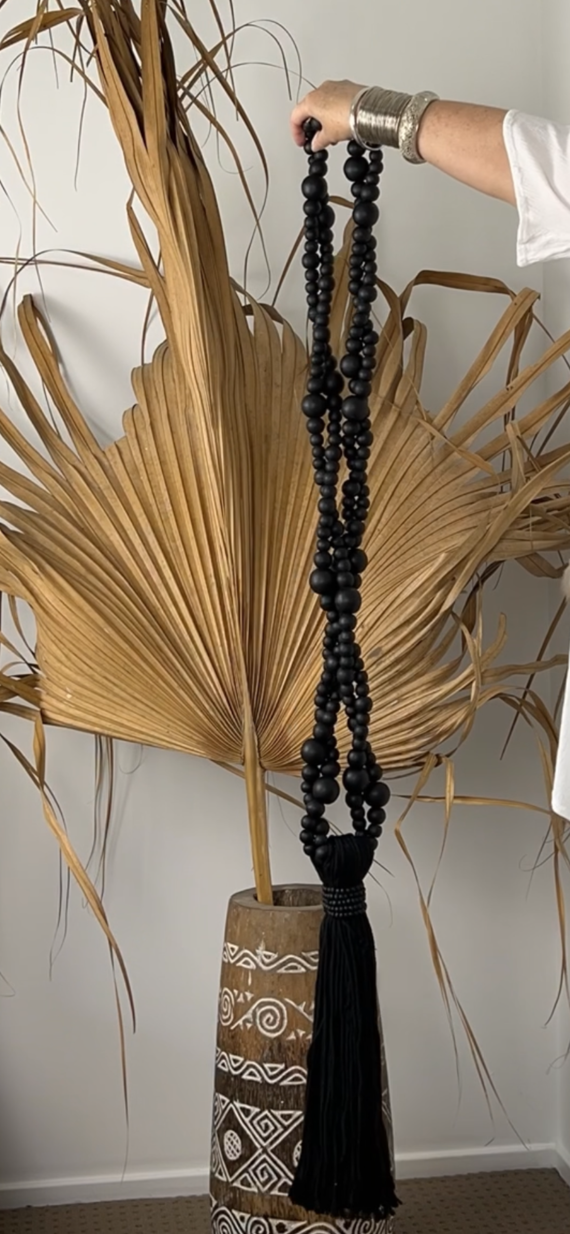 Hand held black beaded twisted tassel shown at full length against a large palm leaf in a pot.