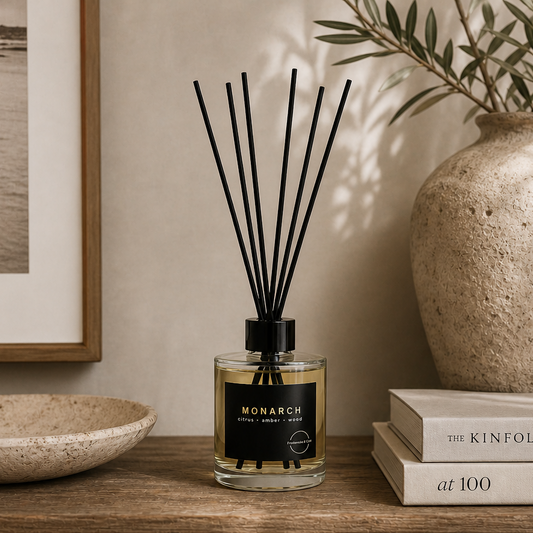 Reed diffuser with black sticks and a clear bottle on a timber shelf and styled with books, plate and a pot in calm beige colouring.