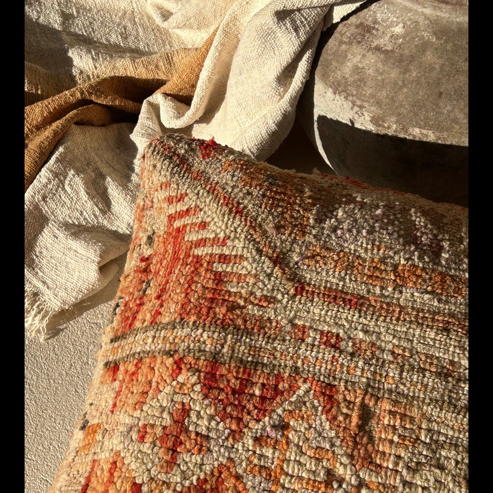 Close up of the corner of the Marrakesh vintage Moroccan floor cushion featuring terracotta, cream and sand tones with geometric tribal patterns, in the sun and against off white flooring next to a pot with a mustard throw.