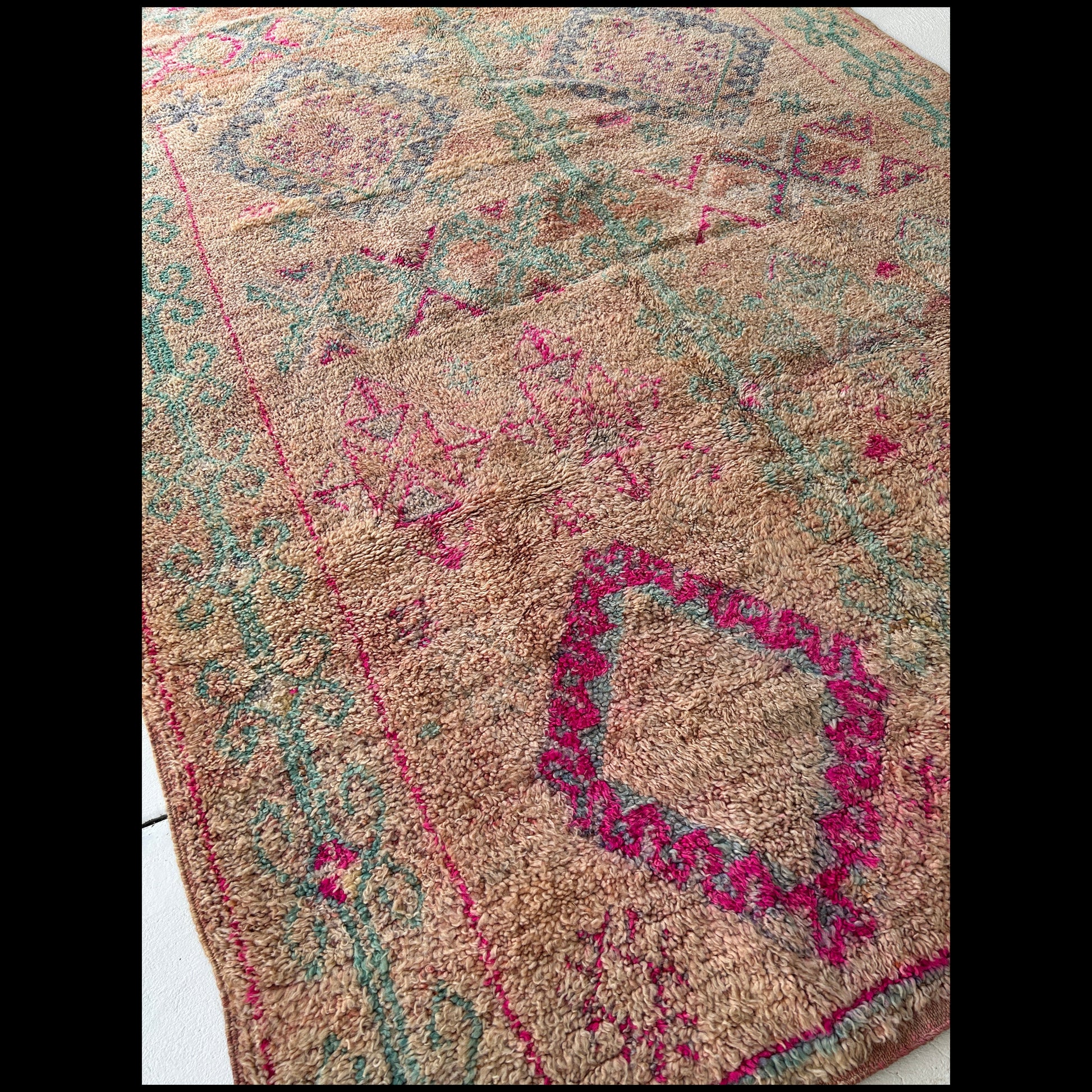 A close up of a Moroccan Bouhssoussen rug with diamond shapes in magenta and teal against a pinky peach background.