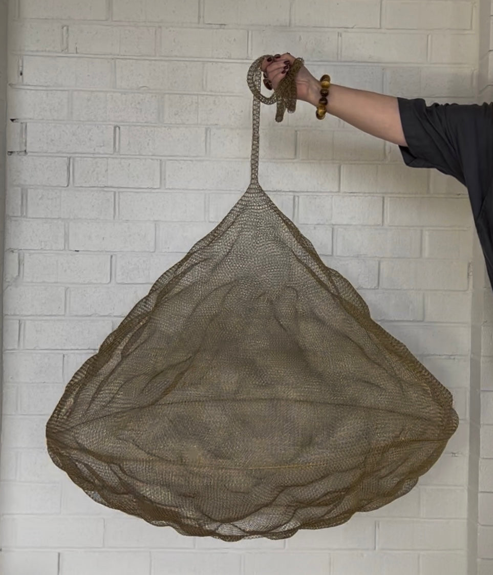 Handwoven Nomad Pendant Brass Mesh Statement Light shown against a white brick wall.
