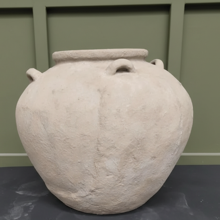 A rustic, light-toned fibre cement urn with a textured, weathered finish and small decorative side handles, displayed against a green panelled wall.