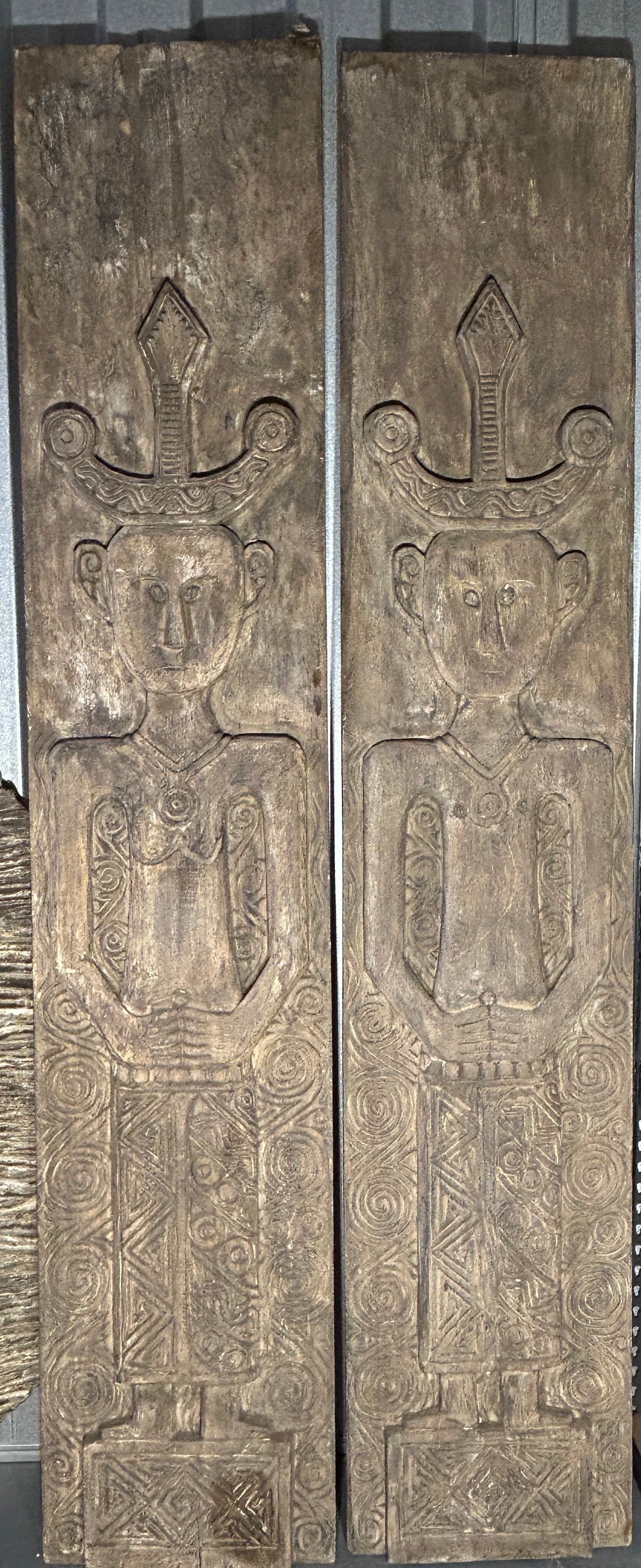 The image is of a pair of hand-carved timber tribal wall panels, known as The Kinship Panels, depicting husband and wife figures with intricate geometric and spiral carvings, ideal for statement foyer or commercial interiors.