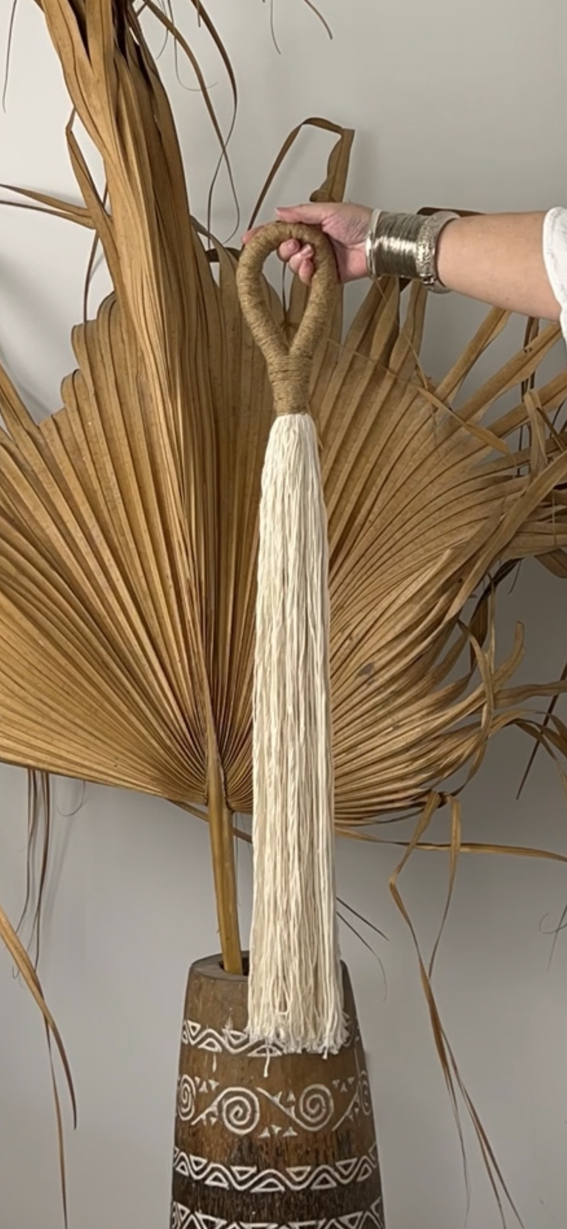 A hand holding a white cotton and jute tassel against the backdrop of a dried palm leaf.