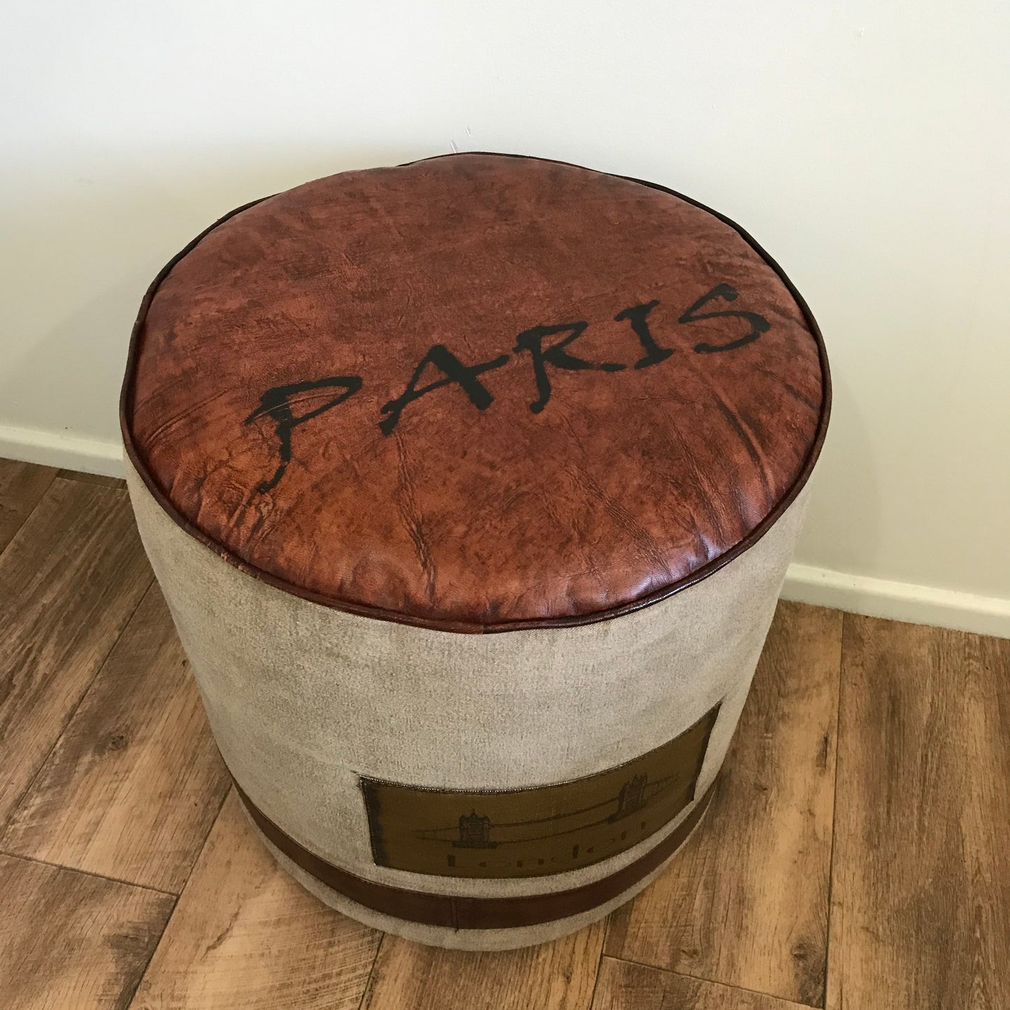 A brown leather ottoman with the word 'PARIS' embossed on top, placed on a wooden floor against a plain wall.